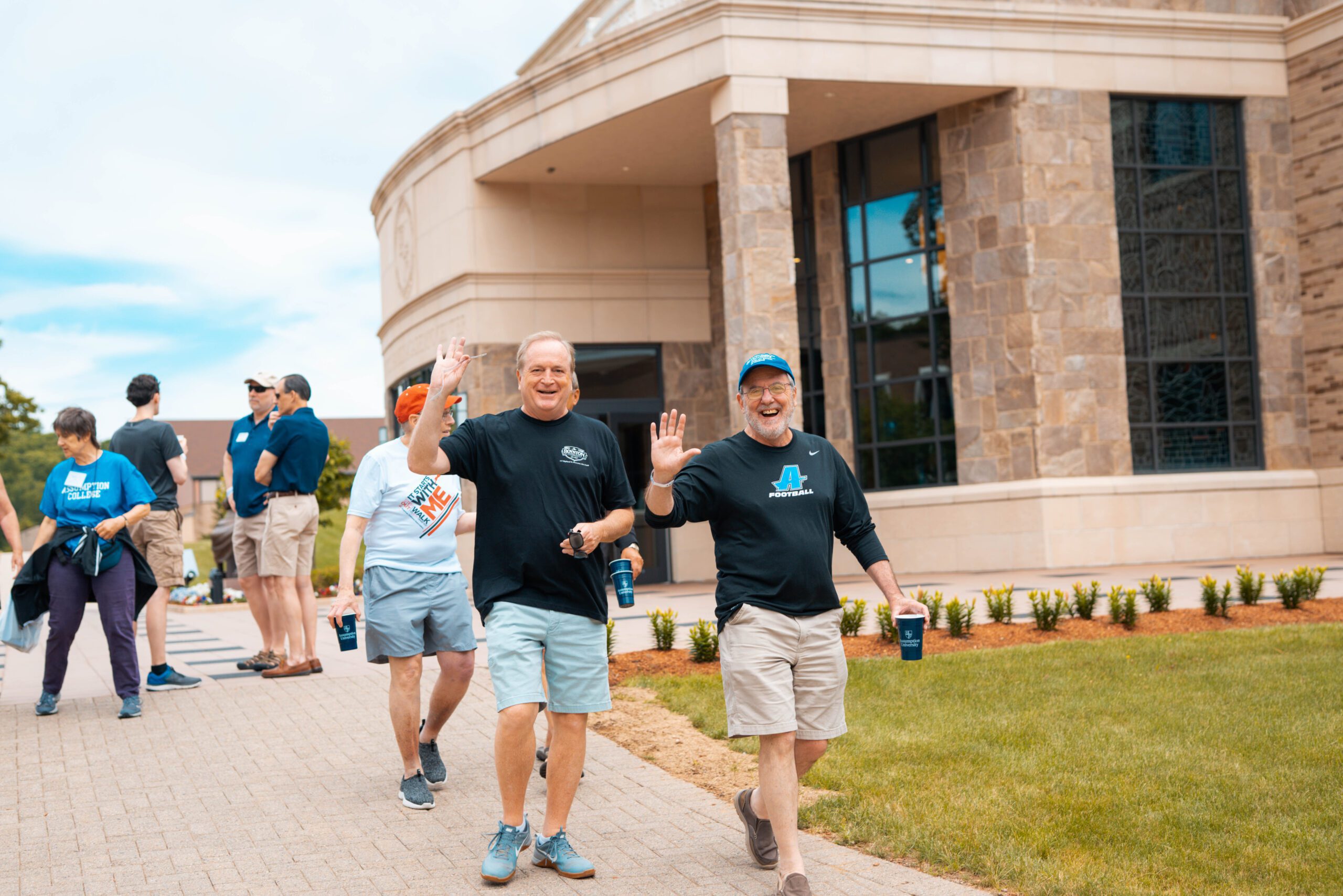 Two male Assumption alumni waving at the camera as they walk past Tsotsis.