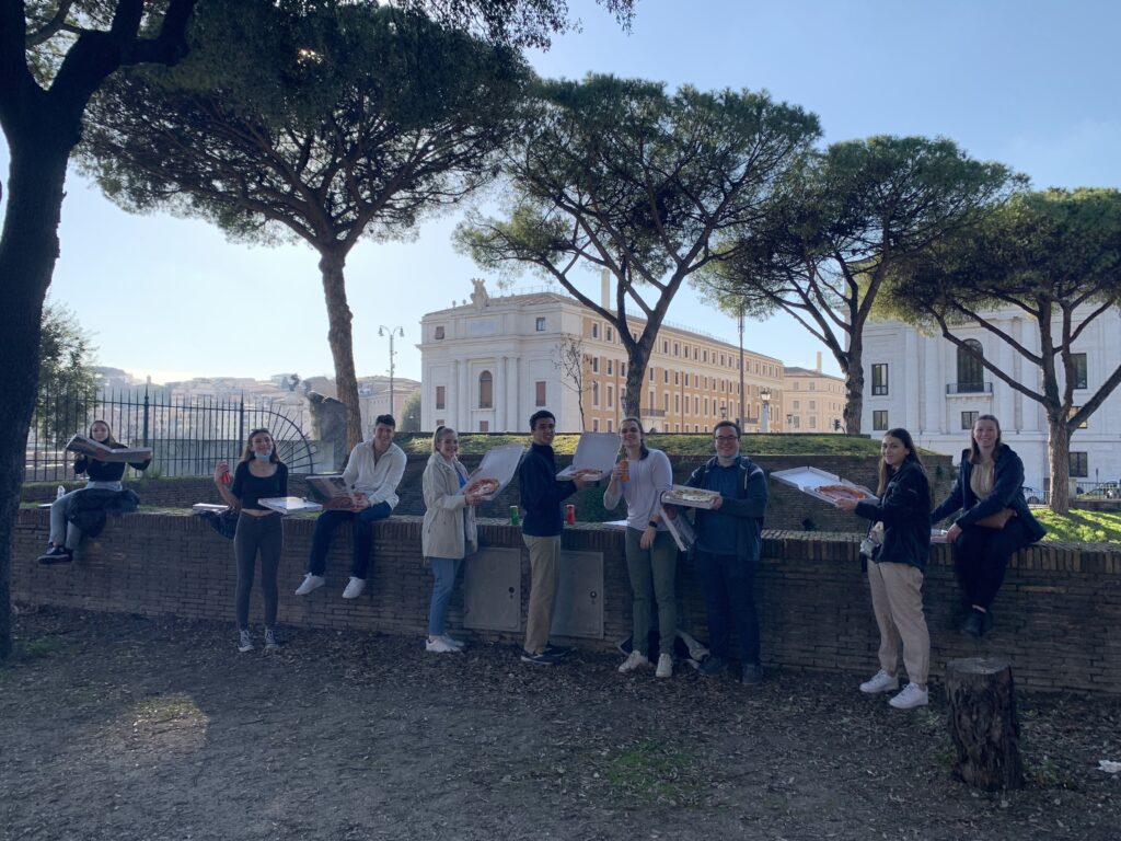Group of students in Rome eating pizza outside along a wall.