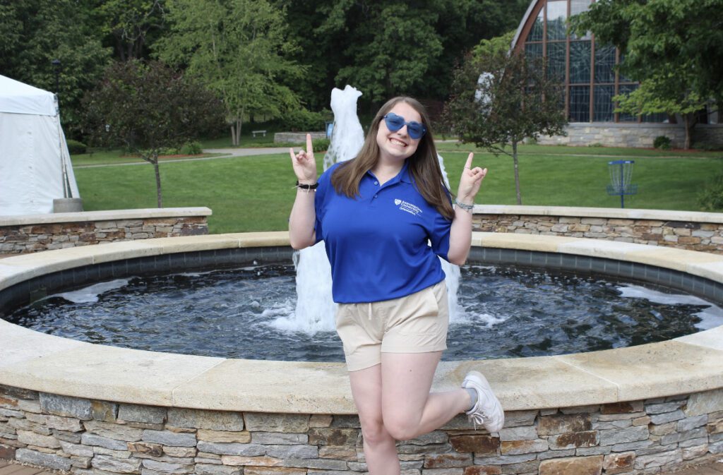 Picture of a female student wearing a blue polo and khaki shorts making a peace sign with her hands.